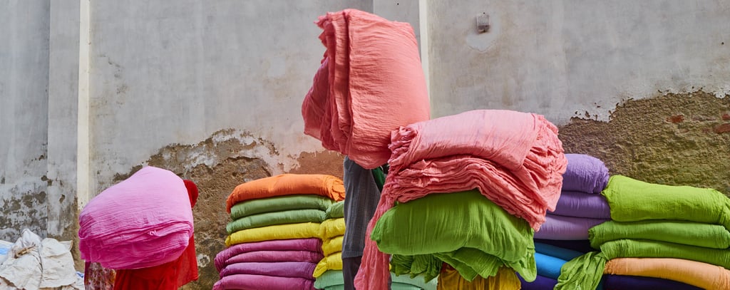 Stacks of brightly colored fabric bundles, including pink, green, orange, purple, and blue, arranged against a weathered concrete wall