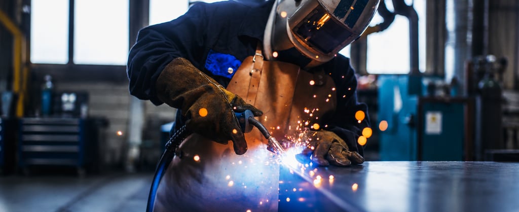 Welder wearing protective equipment working on metal as bright sparks fly in an industrial workshop.