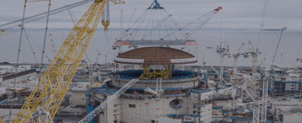 Large crane lifting a reactor dome into place at a coastal nuclear power station construction site.