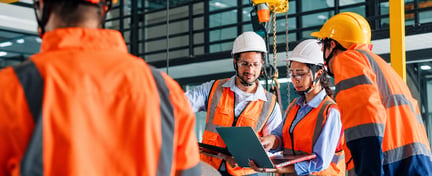 Workers wearing PPE reviewing documents and a laptop in an industrial facility.