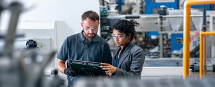 Two professionals wearing safety glasses review information on a tablet in a manufacturing facility.