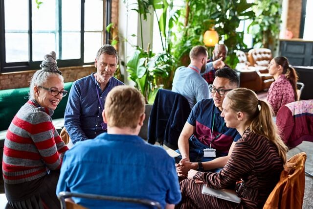 Group of people sitting in a circle in a well-lit indoor space with large windows, plants, and cozy seating, engaged in collaborative discussion or team meeting.